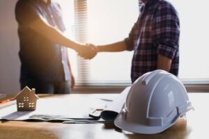 Close-up of two people shaking hands over a desk holding a rolled blueprint, a small wooden house model, and a white hard hat in the foreground.